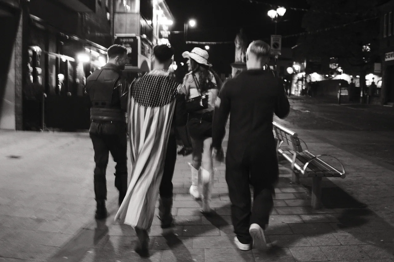 A group walks away through Ormskirk, with a pale cape and a light hat catching the street lighting. Black and white photograph by Mark C Grant.