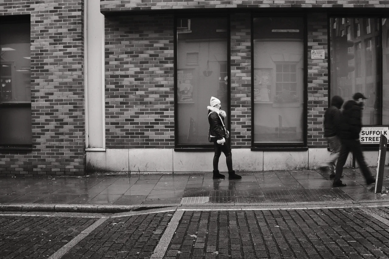 A pedestrian pauses beside tall windows while two blurred figures pass on Suffolk Street, Liverpool. Black and white photograph by Mark C Grant.