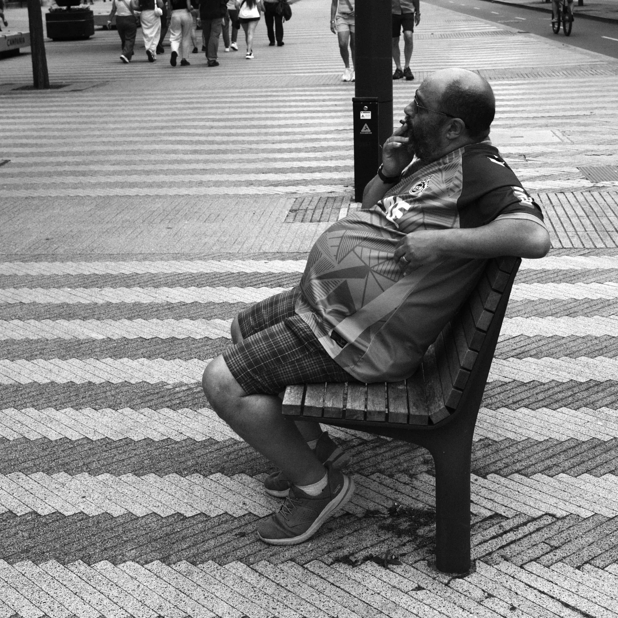 A seated person leaning back on a bench beside a patterned pavement, in black and white. Photograph by Mark C Grant.