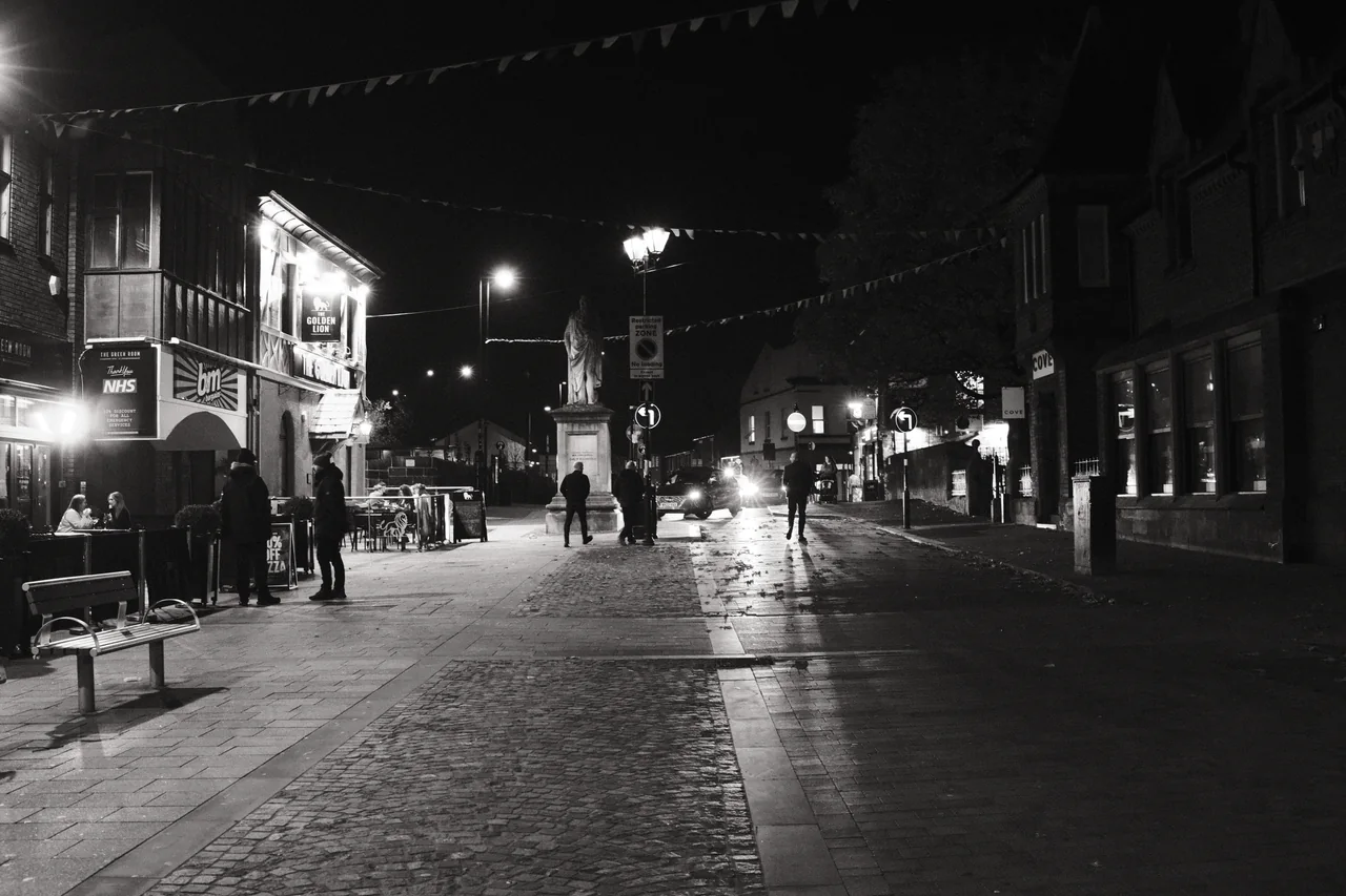Figures stand and walk beneath bunting beside the Golden Lion, with headlights beyond the monument. Black and white photograph by Mark C Grant.