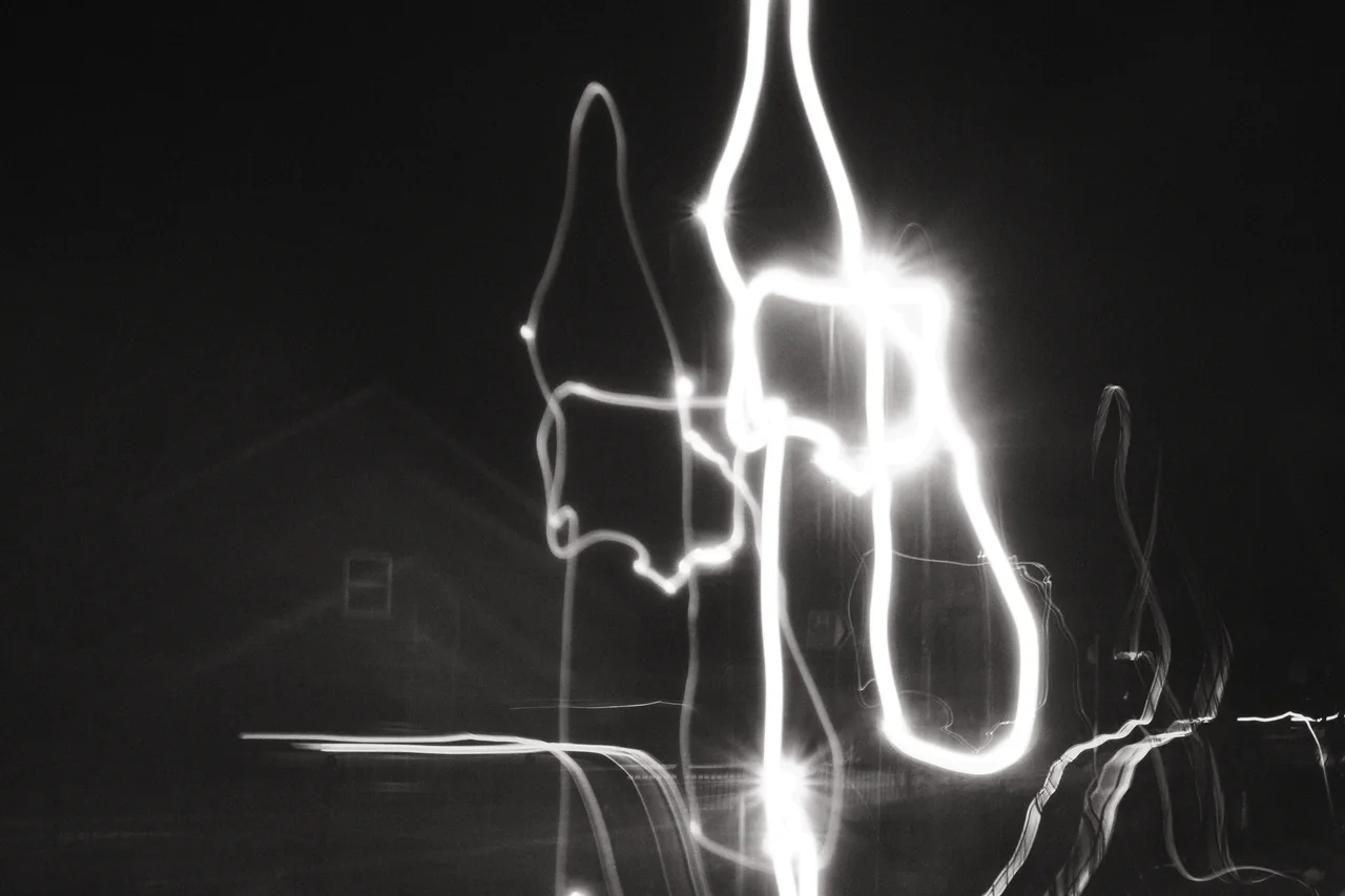 Long white light trails loop across a night scene, with the faint outline of a house behind them. Black and white photograph by Mark C Grant.