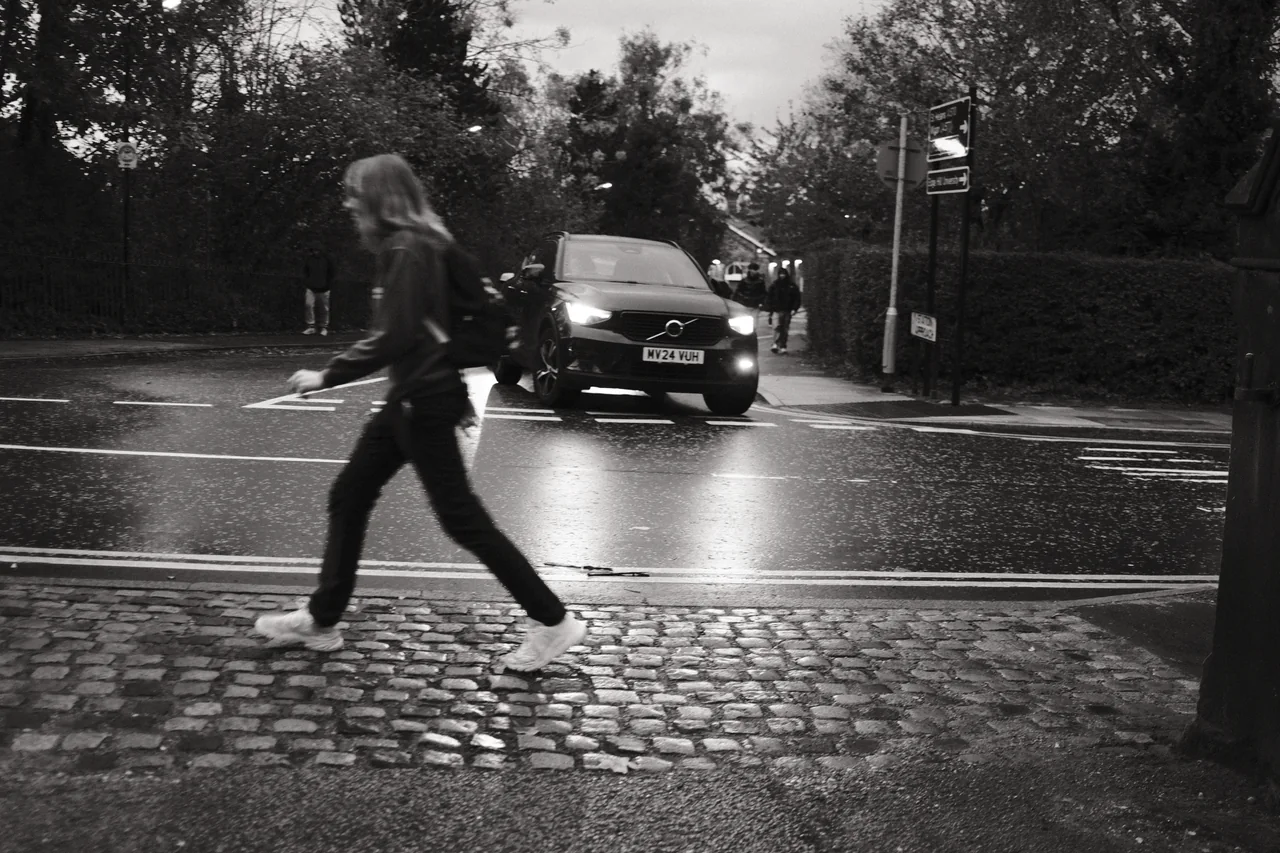A pedestrian strides across the foreground beside a wet road in Ormskirk as headlights reflect behind them. Black and white photograph by Mark C Grant.