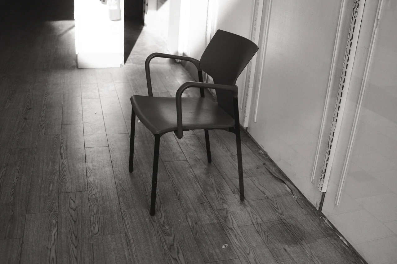 An empty chair sits beside an open doorway, with pale walls and diagonal floorboards around it. Black and white photograph by Mark C Grant.