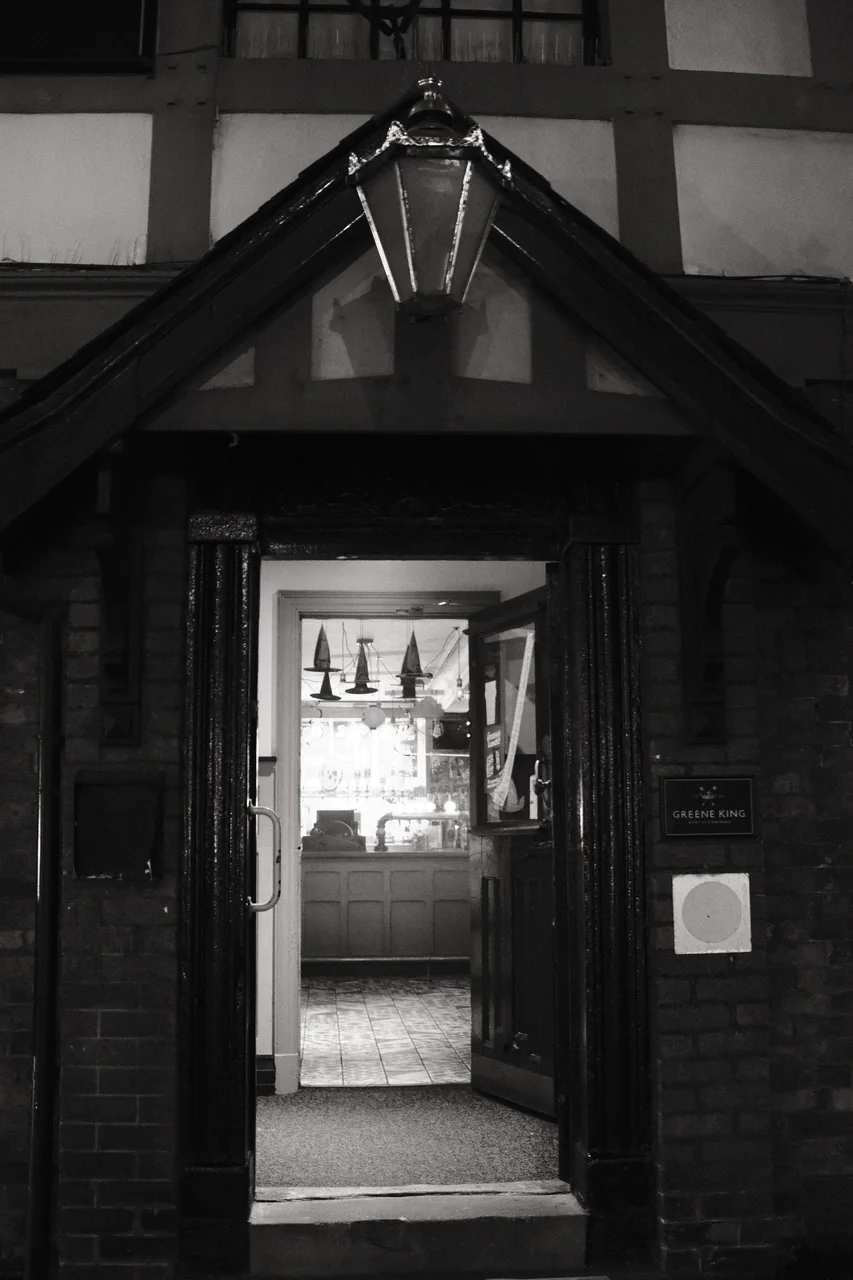 An open pub door reveals a bright interior and hanging pointed hats beneath a dark timber porch. Black and white photograph by Mark C Grant.
