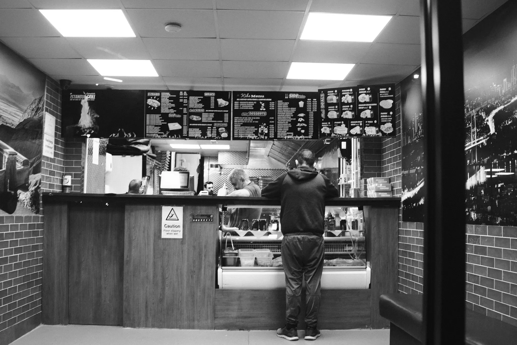 A person standing at an illuminated takeaway counter. Black and white photograph by Mark C Grant.