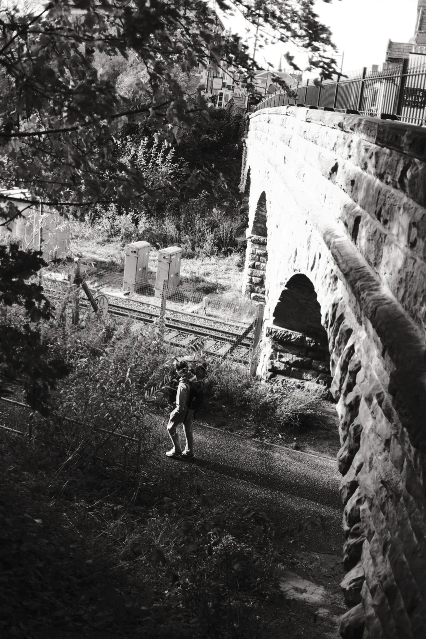 A lone figure stands on the path below a sunlit stone railway bridge, surrounded by foliage, rails and shadow. Black and white photograph by Mark C Grant.