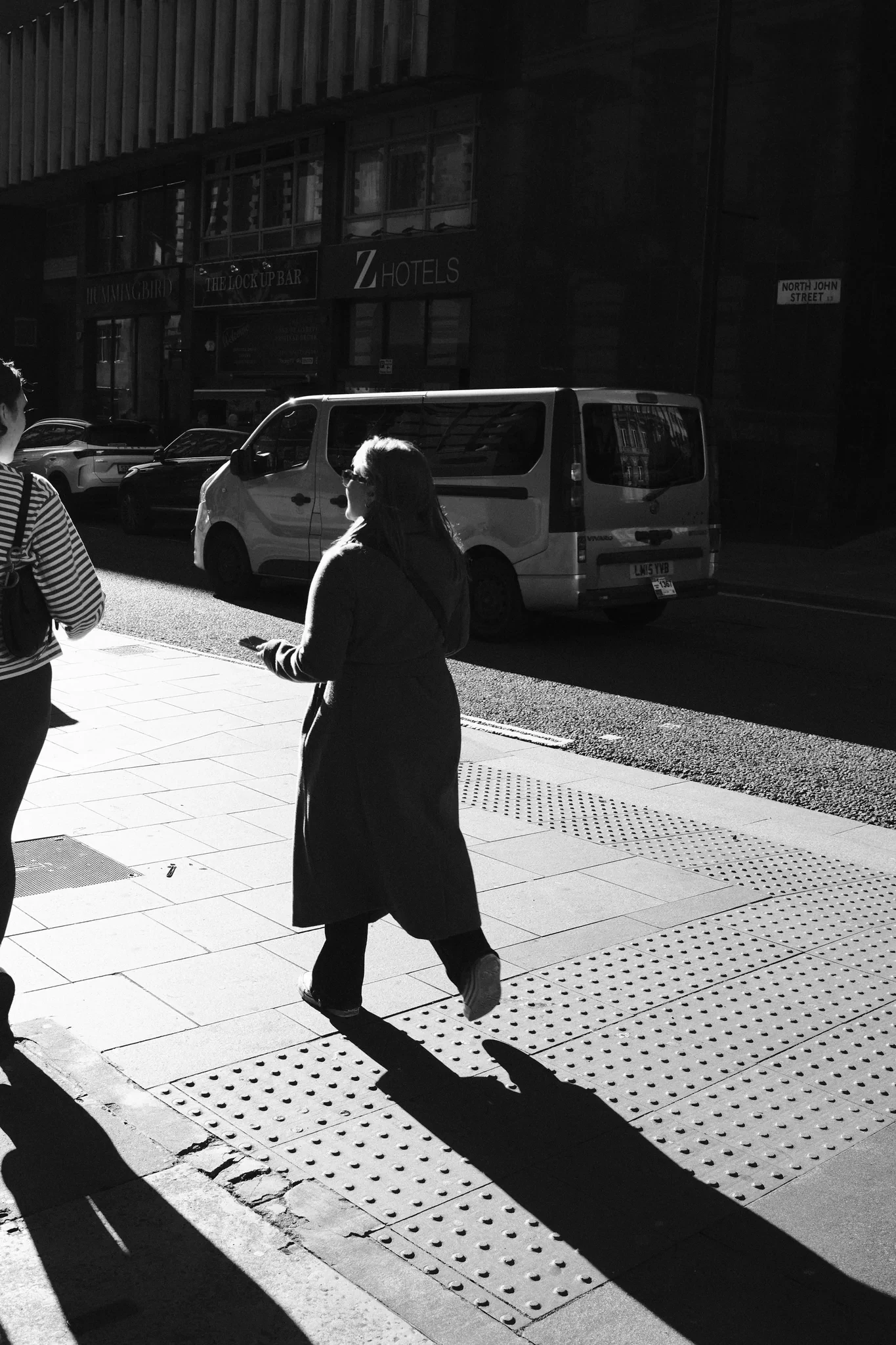 A pedestrian and a long shadow cross a bright patch of pavement in Liverpool city centre. Black and white photograph by Mark C Grant.
