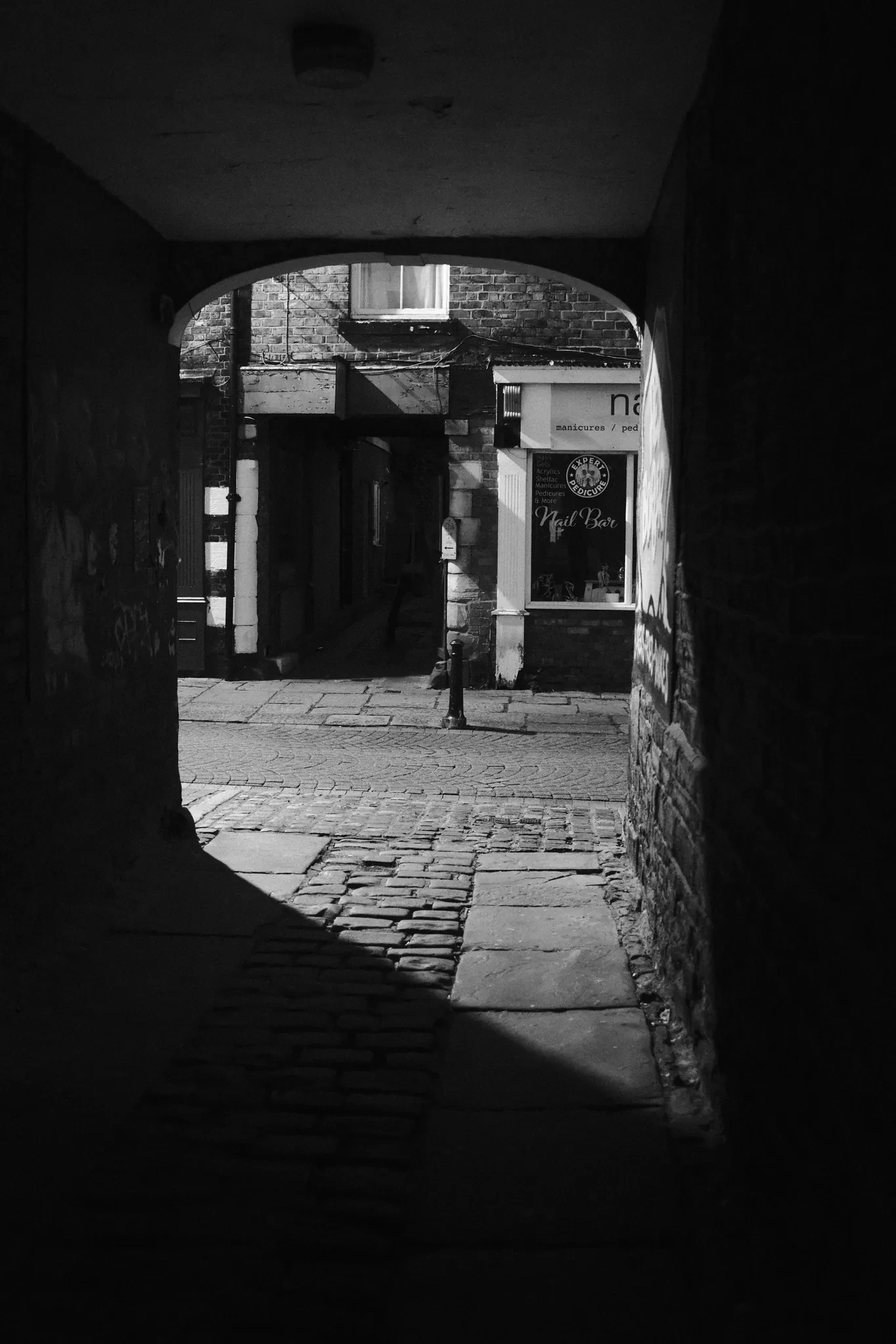 A shop doorway seen from the darkness of a narrow passage off Burscough Street, Ormskirk. Black and white photograph by Mark C Grant.