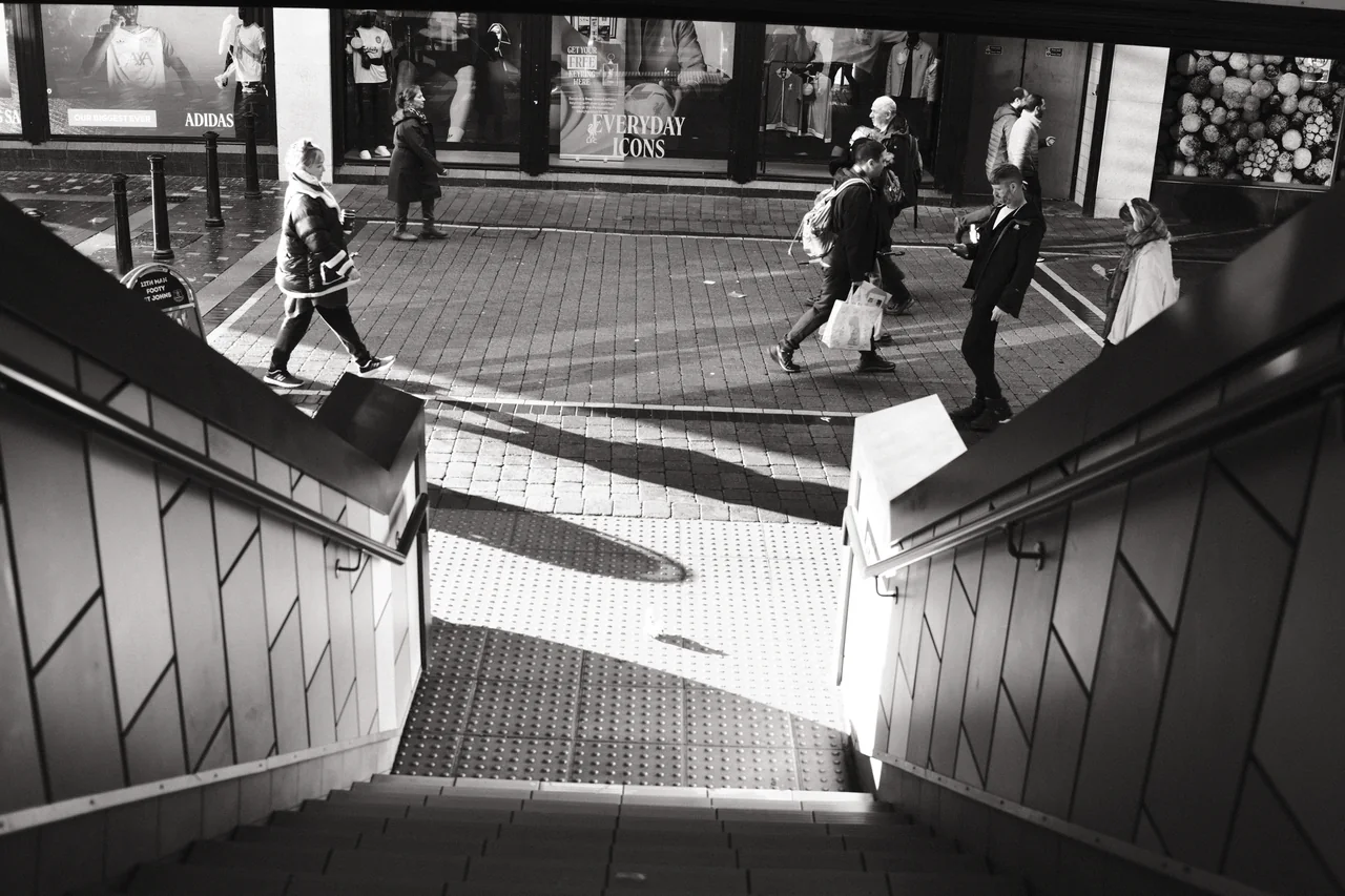 Handrails frame a view down the steps at St Johns Shopping Centre, where pedestrians pass through a patch of sunlight. Black and white photograph by Mark C Grant.