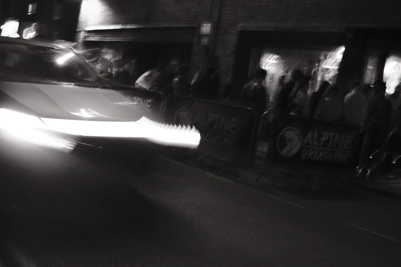 A passing car stretches bright headlight traces across the foreground outside Alpine Ormskirk. Black and white photograph by Mark C Grant.