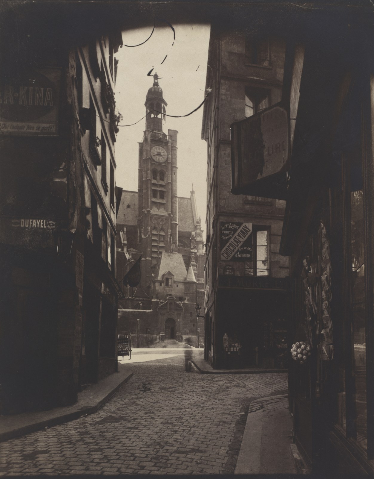 A narrow cobbled Paris street in deep foreground shadow opens towards a sunlit church tower, with shop signs projecting into the view.