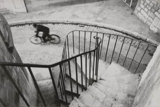 View down a curved staircase and railings towards a blurred cyclist in the street below.