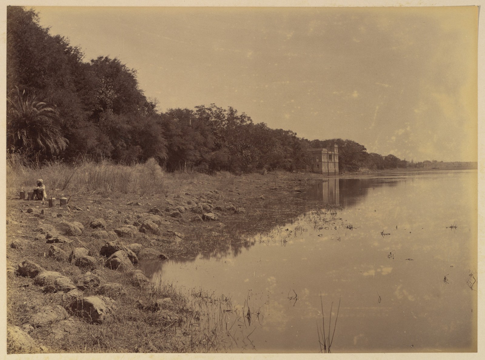 A rocky, planted shoreline curves towards a small distant structure beside calm water, with trees on the left and a broad pale sky.