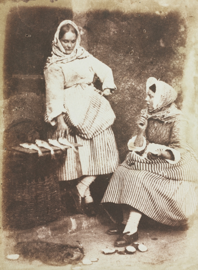 Jeanie Wilson and Annie Linton, one standing and one seated, wear patterned skirts beside a basket holding fish; shells lie at their feet.