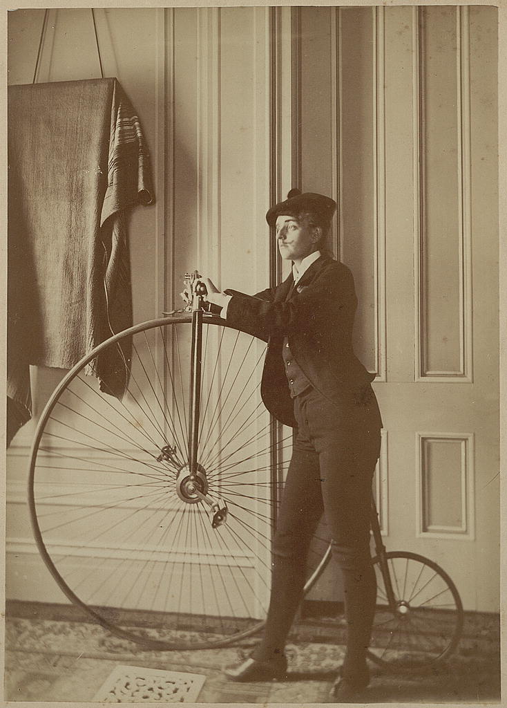 Frances Benjamin Johnston wears a suit, hat and false moustache, standing beside a penny-farthing bicycle in a panelled room.