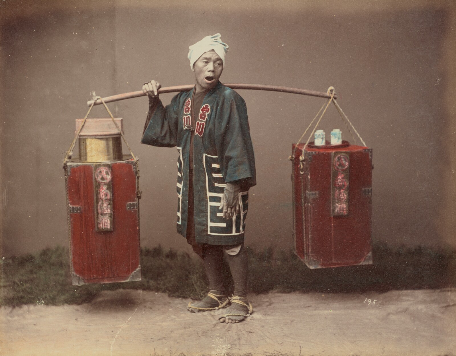 A posed amazake seller stands before a plain studio backdrop, carrying two red boxes suspended from a pole across his shoulder; the photograph is hand-coloured.