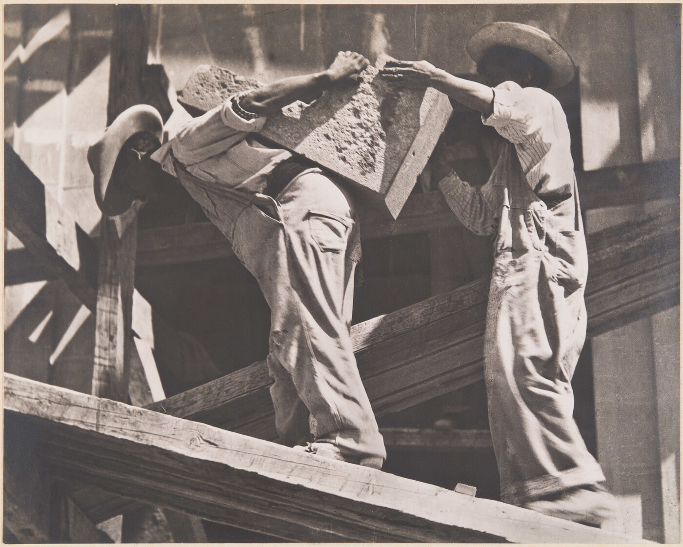 Two workers handle a rough block of stone among wooden beams. The man on the left bends beneath the block while the man at right reaches towards it; their faces are shaded by hats.