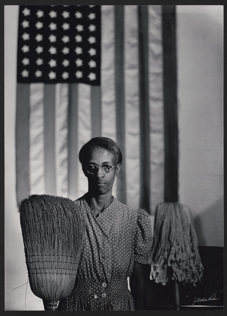 Ella Watson stands in a dotted dress with broom and mop before a large American flag; the flag is softer than the face and broom.
