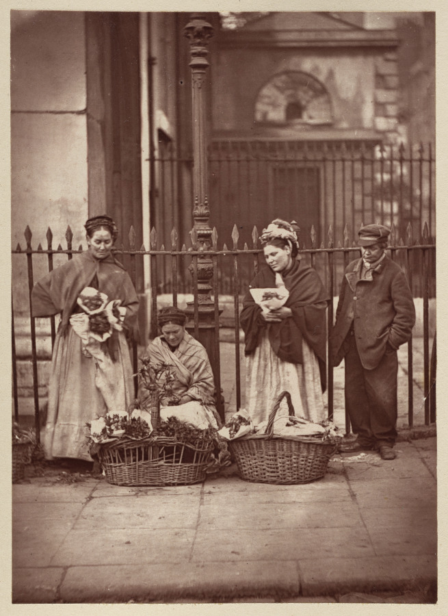 Three flower sellers and a man stand or sit beside large baskets against iron railings; one woman cradles a small child.