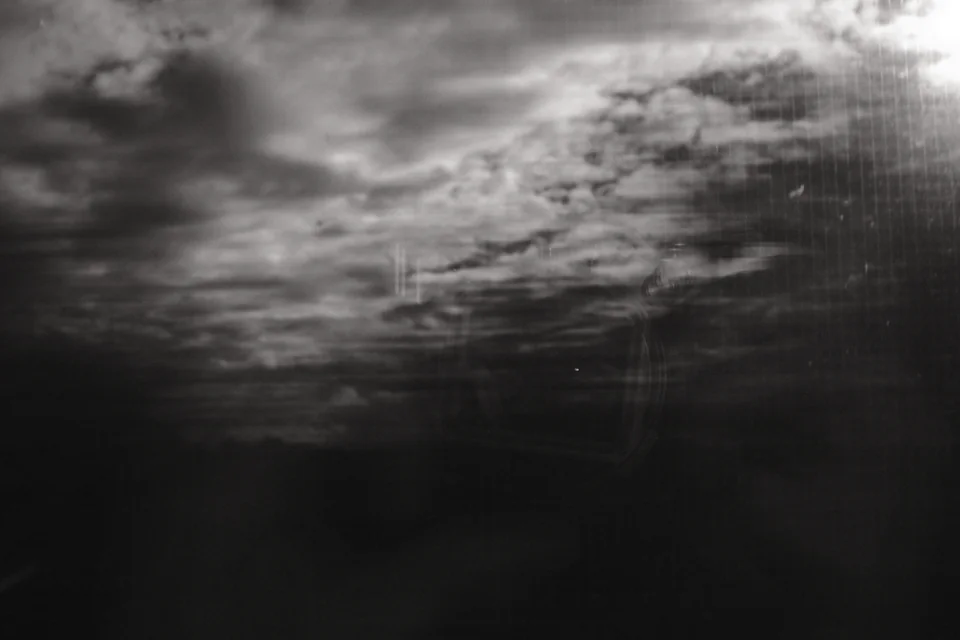 Landscape and clouds seen through a train window, with streaked foreground and the faint reflection of Mark C Grant holding his camera. Black and white photograph by Mark C Grant.