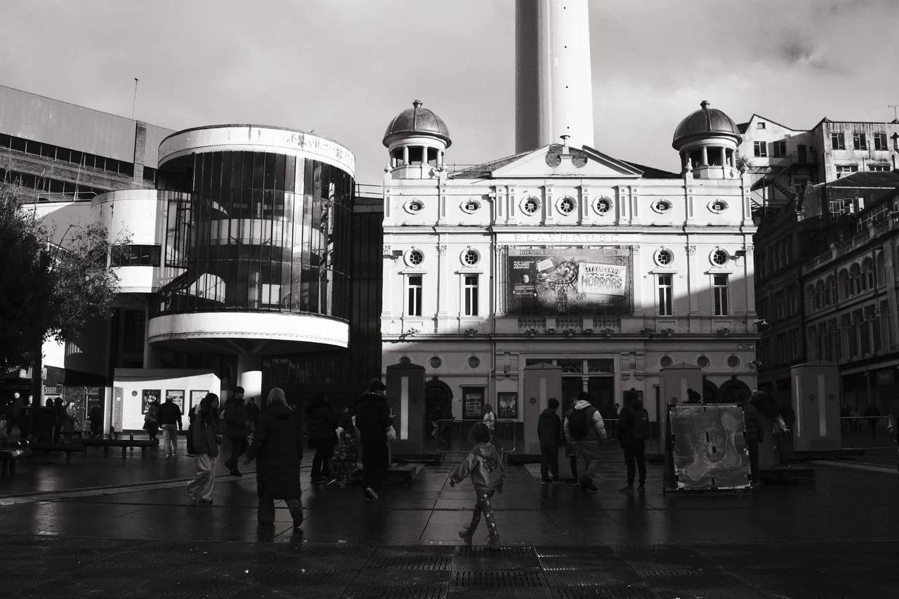Sunlight falls across the Playhouse Theatre façade while pedestrians cross the darker paving of Williamson Square. Black and white photograph by Mark C Grant.
