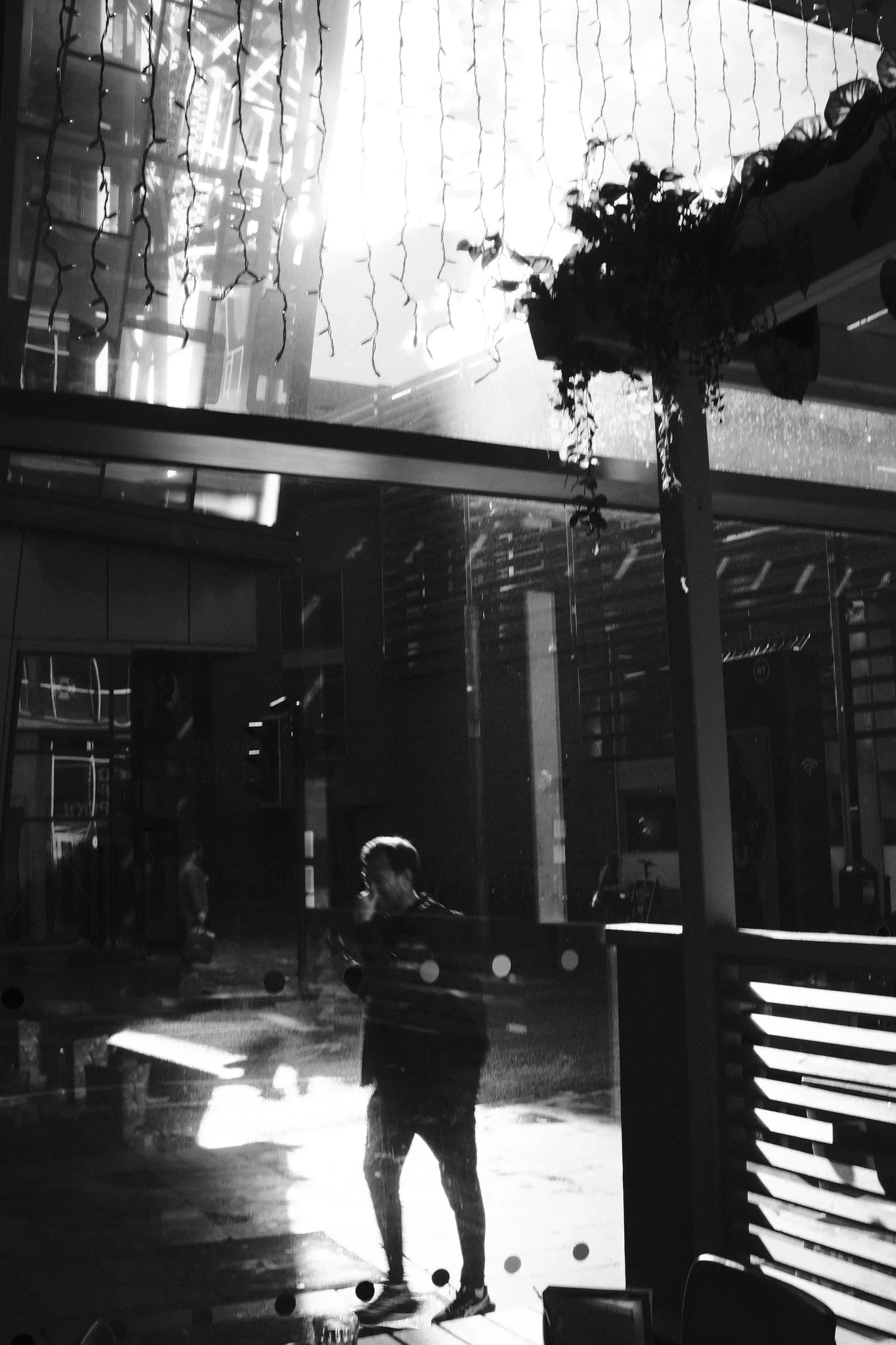 A figure crosses a bright patch of wet pavement beyond a shaded cafe frontage. Black and white photograph by Mark C Grant.