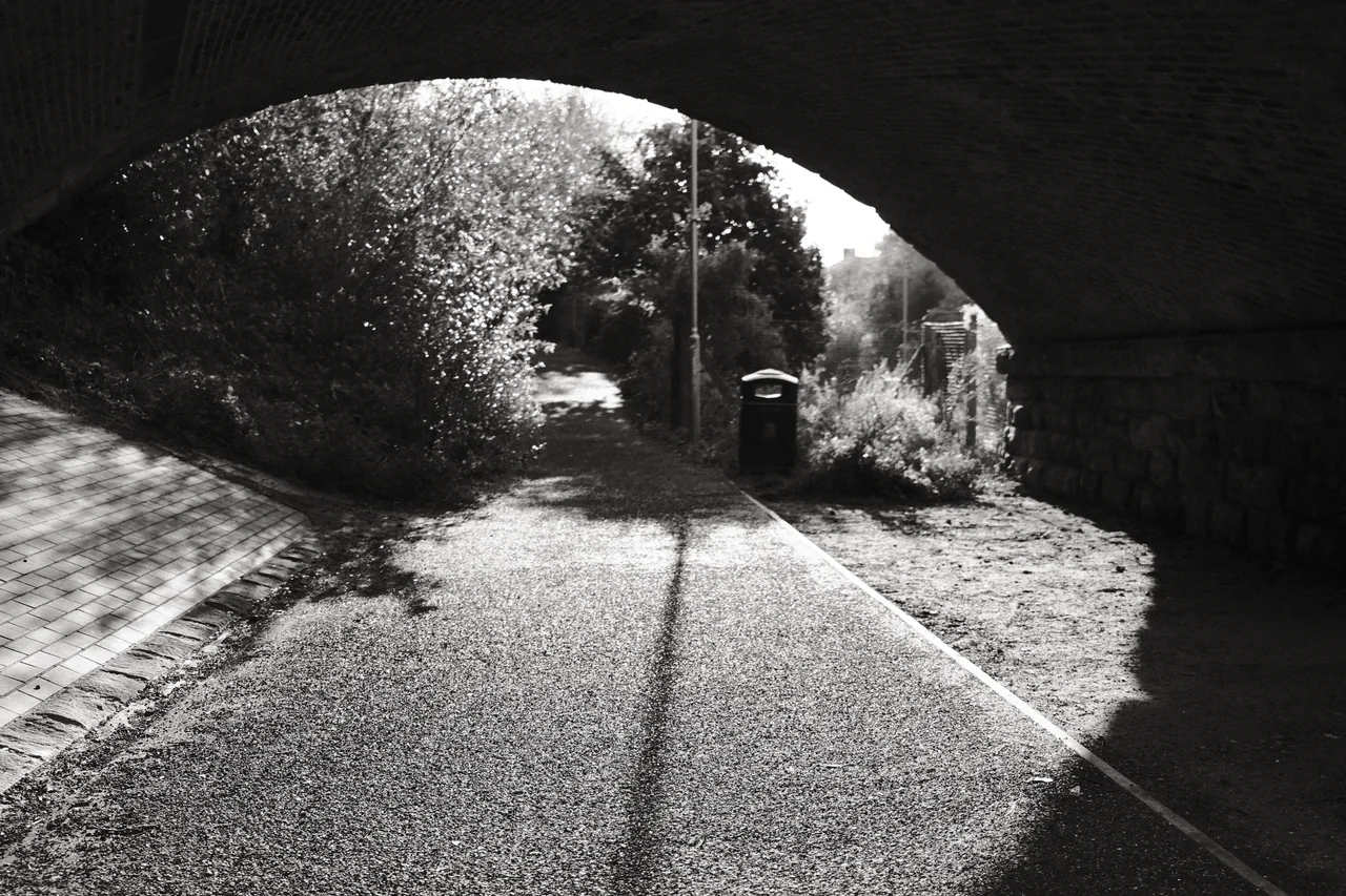 A dark bridge arch frames a sunlit path, a litter bin and a long lamp-post shadow. Black and white photograph by Mark C Grant.