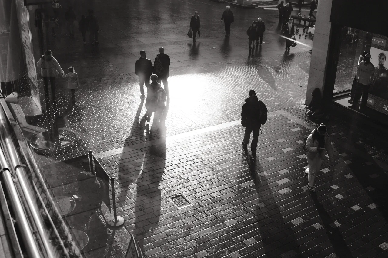 Seen from above, pedestrians cast long shadows across the wet paving of Williamson Square in Liverpool. Black and white photograph by Mark C Grant.