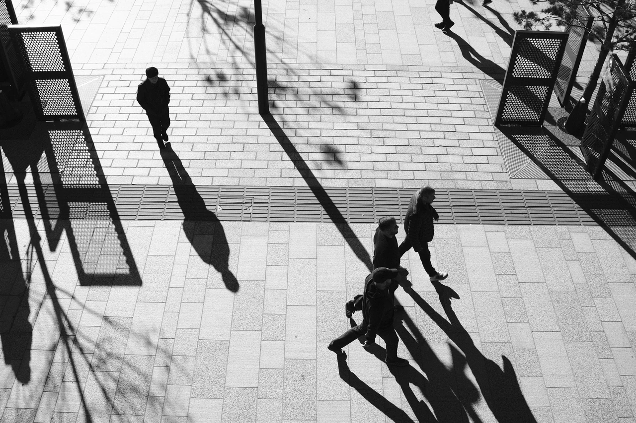 Viewed from above, visitors and their long shadows cross the paving outside Bramley Moore (Hill Dickinson), Liverpool. Black and white photograph by Mark C Grant.