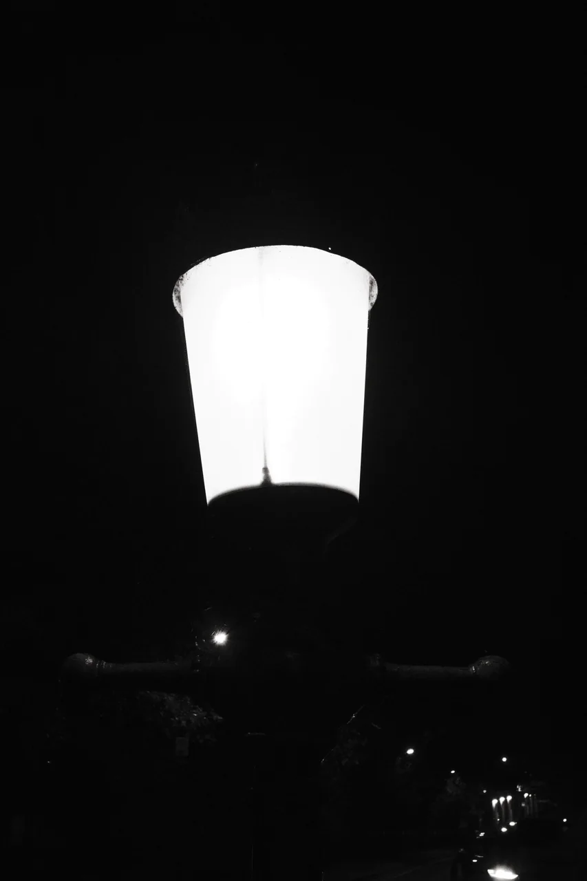 A bright street lamp fills the upper part of a vertical frame, with smaller lights receding into darkness below. Black and white photograph by Mark C Grant.