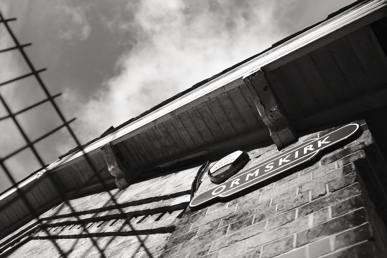 The Ormskirk station sign and roofline run diagonally across the frame, behind a blurred foreground fence. Black and white photograph by Mark C Grant.