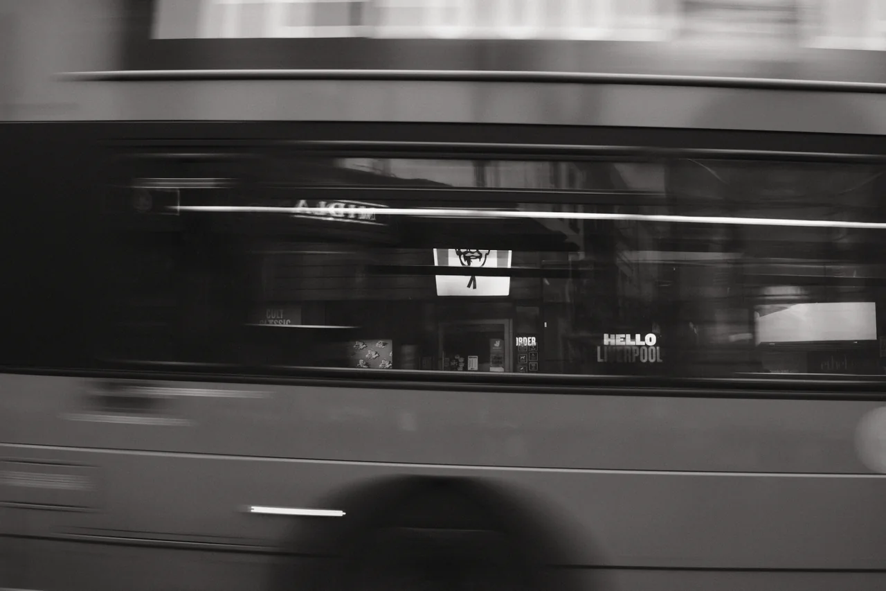 A passing bus blurs across Liverpool shopfronts, layering dark windows, lettering and horizontal streaks. Black and white photograph by Mark C Grant.