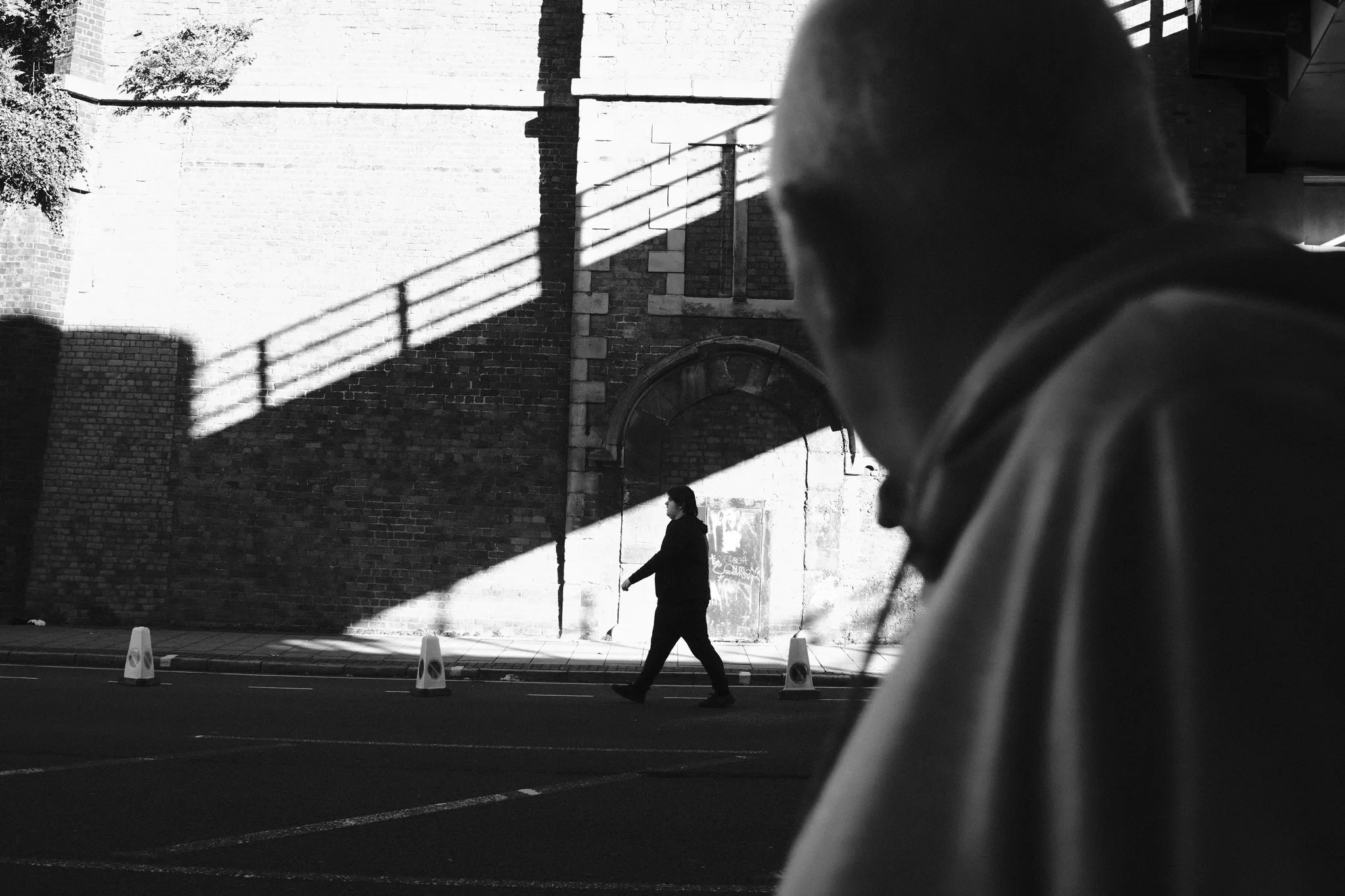 A passer-by fills the foreground while another figure crosses a wedge of sunlight beneath a brick stairway in Liverpool. Black and white photograph by Mark C Grant.
