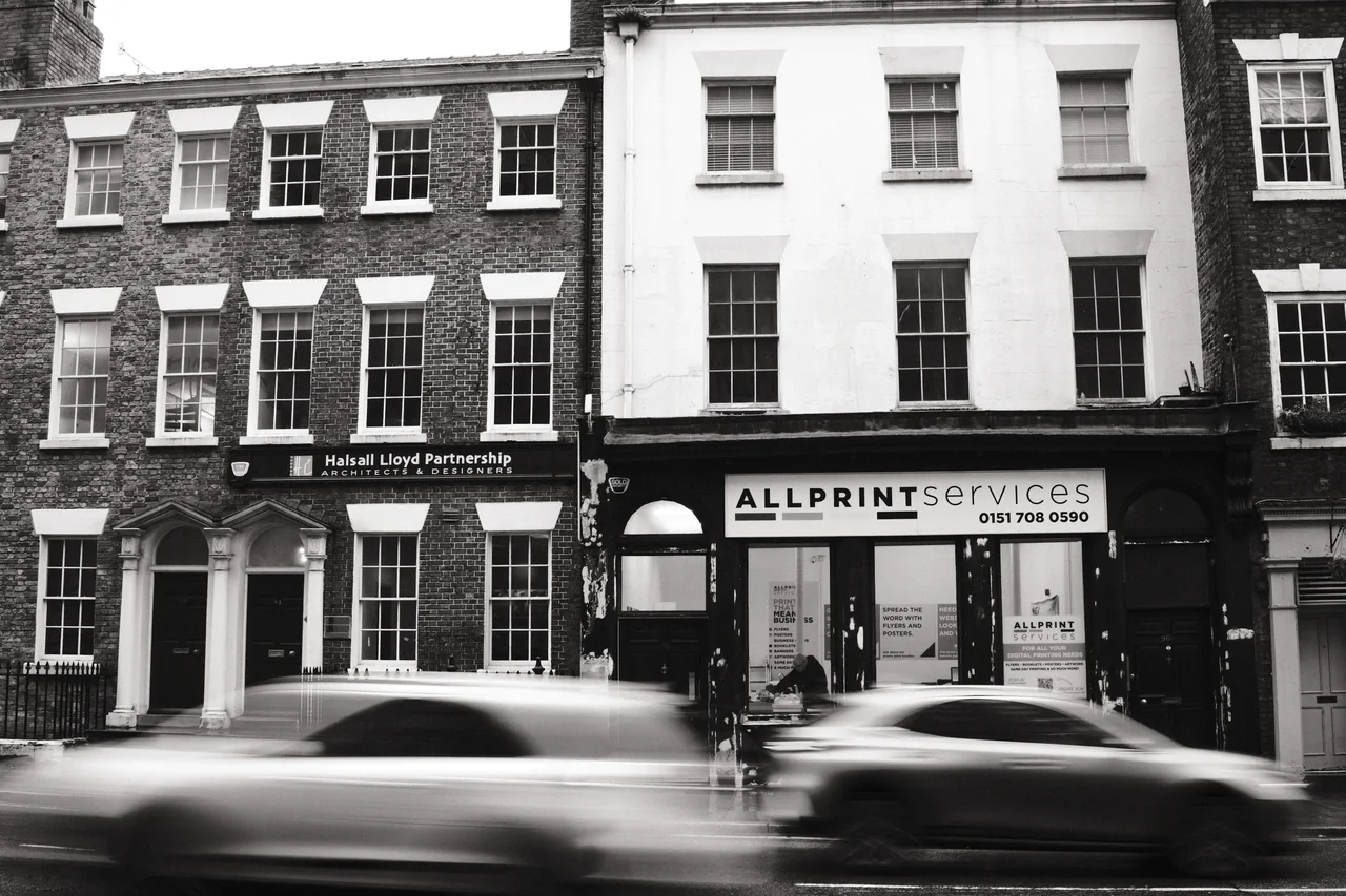 Two cars blur across the Georgian shopfronts beside Allprint Services on Duke Street, Liverpool. Black and white photograph by Mark C Grant.