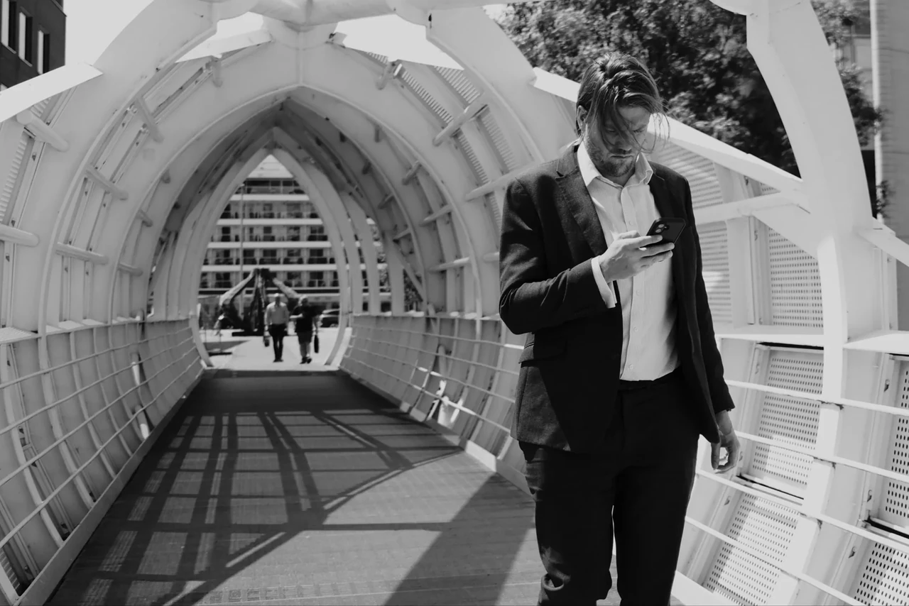 A person checks a phone beneath the repeating white arches of Princes Dock footbridge. Black and white photograph by Mark C Grant.
