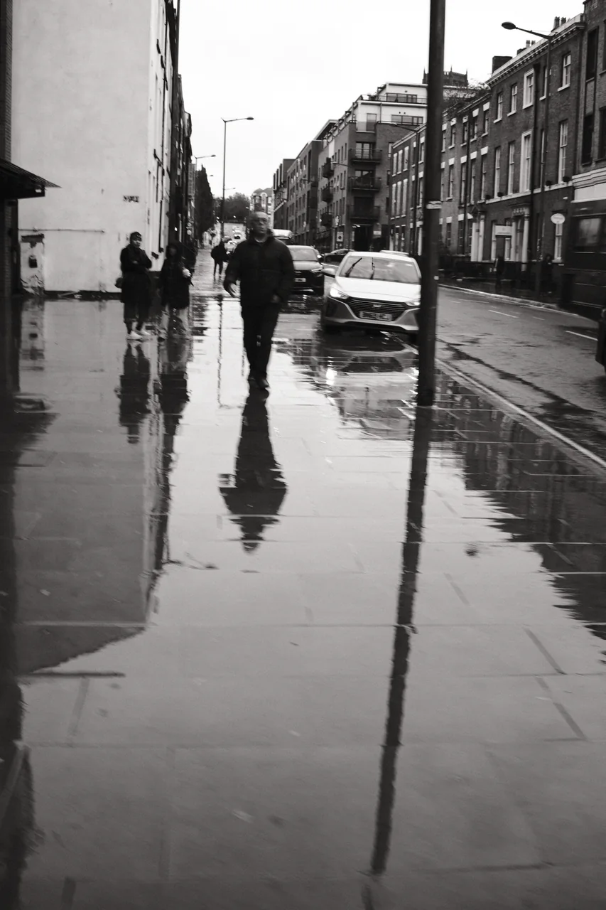 Pedestrians and a lamp post are reflected in rainwater along Duke Street in Liverpool. Black and white photograph by Mark C Grant.