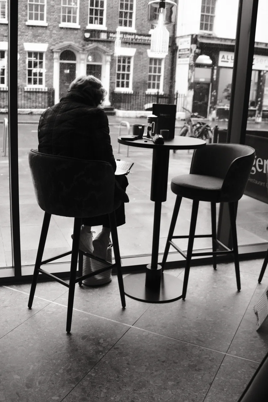 A seated customer reads beside the window at Voyagers, with Duke Street shopfronts beyond the glass. Black and white photograph by Mark C Grant.