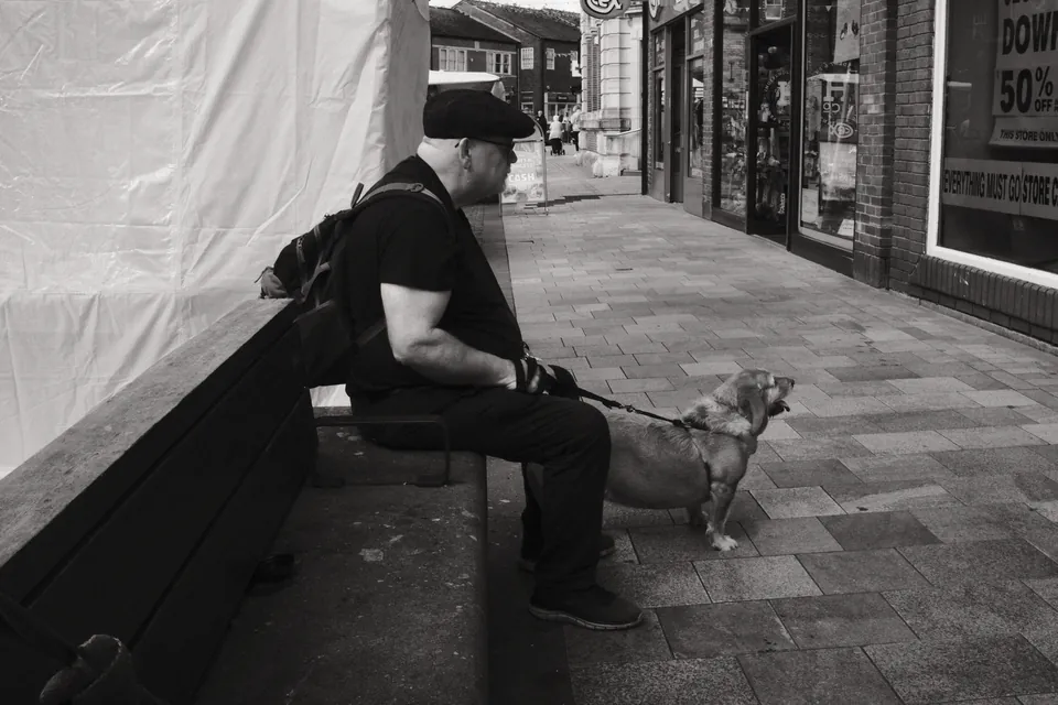 A person and a small dog pause beside the shops. Black and white photograph by Mark C Grant.