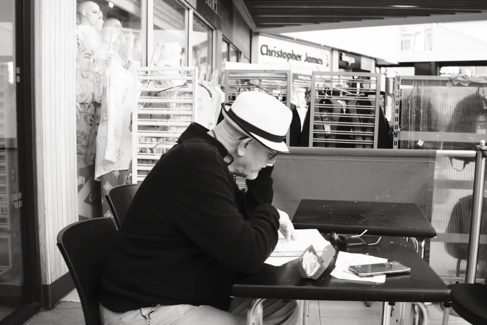 A person in a light hat sits at a café table in a covered shopping space. Black and white photograph by Mark C Grant.
