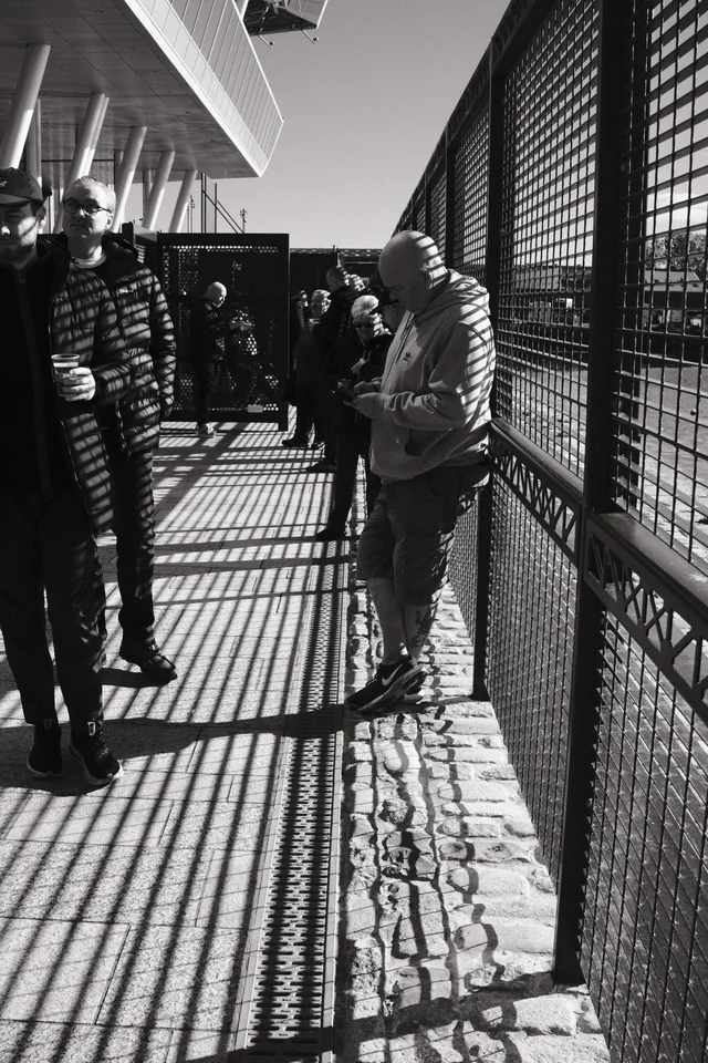 A man looks down beside a railing while a grid of sunlight and shadow crosses the walkway outside Everton’s stadium. Black and white photograph by Mark C Grant.