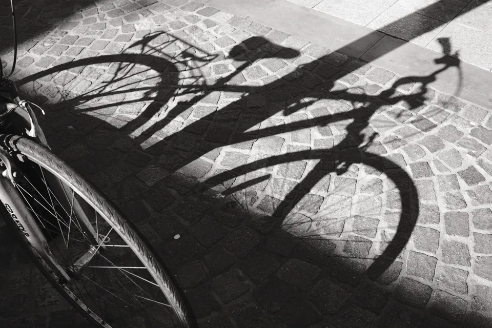The wheels and frame of a bicycle cast overlapping shadows across cobbles. Black and white photograph by Mark C Grant.