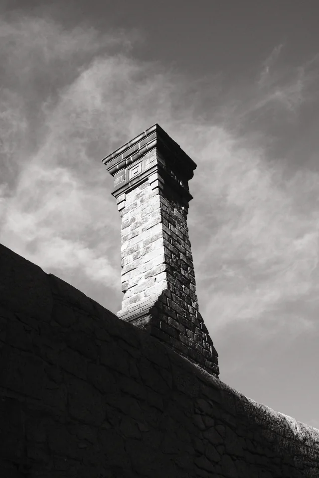 A brick chimney rises above a dark roofline into a streaked sky on Liverpool’s dockside. Black and white photograph by Mark C Grant.