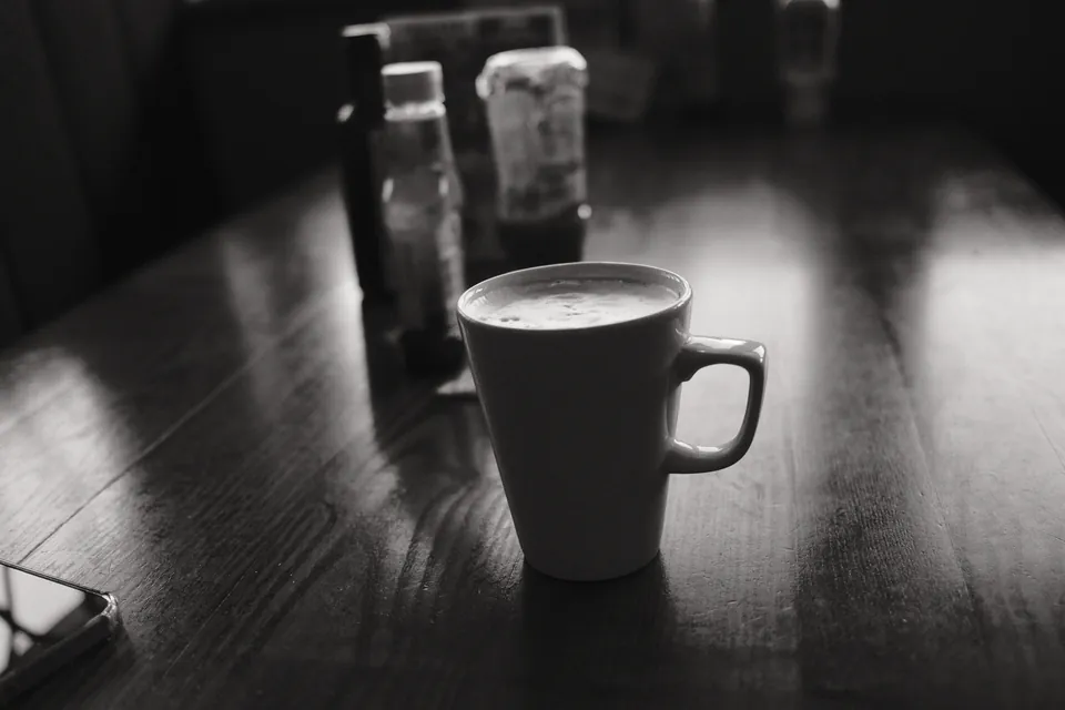 A cup of coffee rests on a dark table beside soft reflected light. Black and white photograph by Mark C Grant.