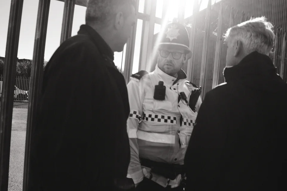 Two men speak with a police officer as low sunlight floods the street near Everton’s stadium. Black and white photograph by Mark C Grant.