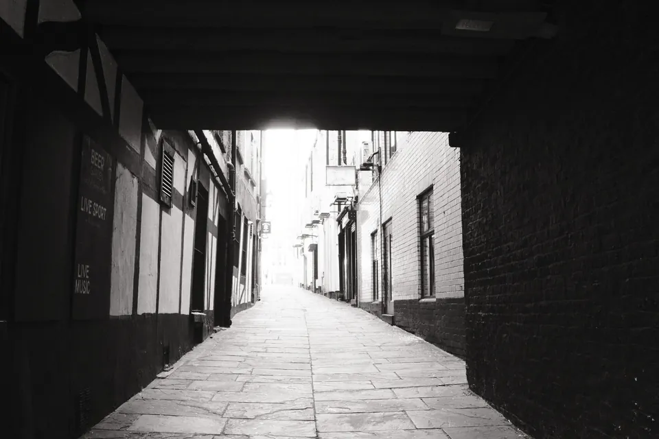 A narrow paved lane emerging from the shadow of an overhead structure. Black and white photograph by Mark C Grant.