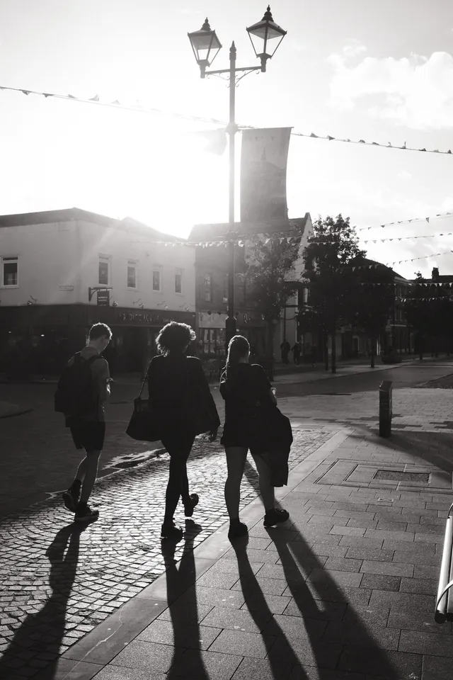Three pedestrians cast long shadows across the paving in Ormskirk town centre. Black and white photograph by Mark C Grant.
