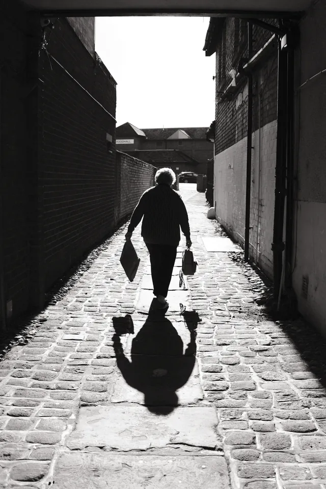 A figure carrying bags walks through a sunlit passage, casting a long shadow over the cobbles. Black and white photograph by Mark C Grant.