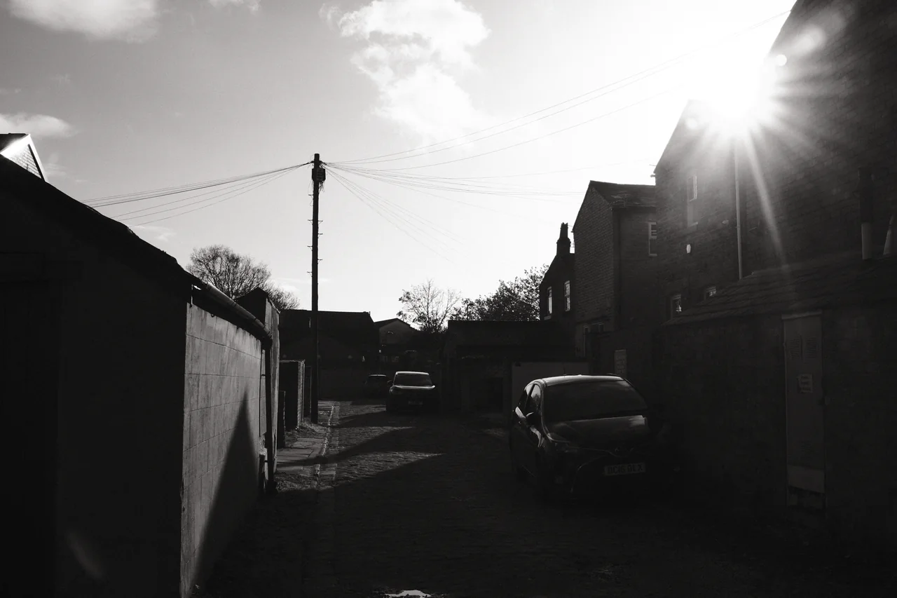 Sunlight breaks over a roof above a shadowed backstreet, parked cars and overhead wires. Black and white photograph by Mark C Grant.