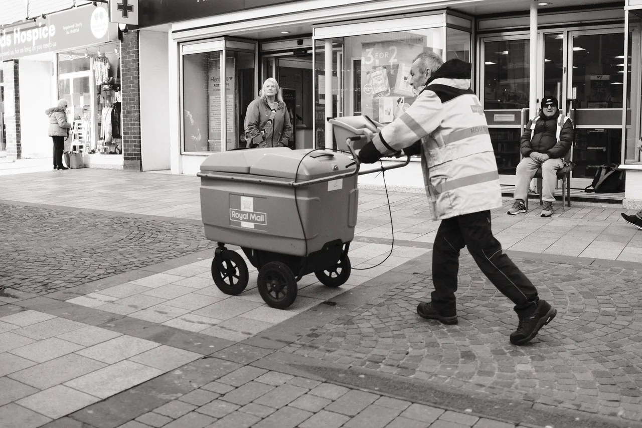 A postal worker pushes a Royal Mail trolley past people outside a row of shops. Black and white photograph by Mark C Grant.
