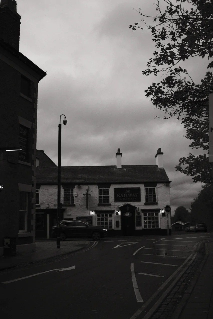 The Railway pub on Derby Street in Ormskirk sits beneath an overcast sky, framed by a dark building and autumn branches. Black and white photograph by Mark C Grant.